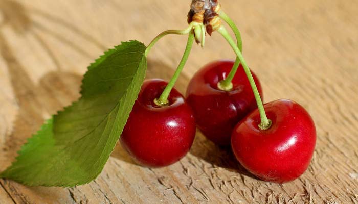 Tres cerezas rojas con el rabillo y una hoja sobre una tabla de madera
